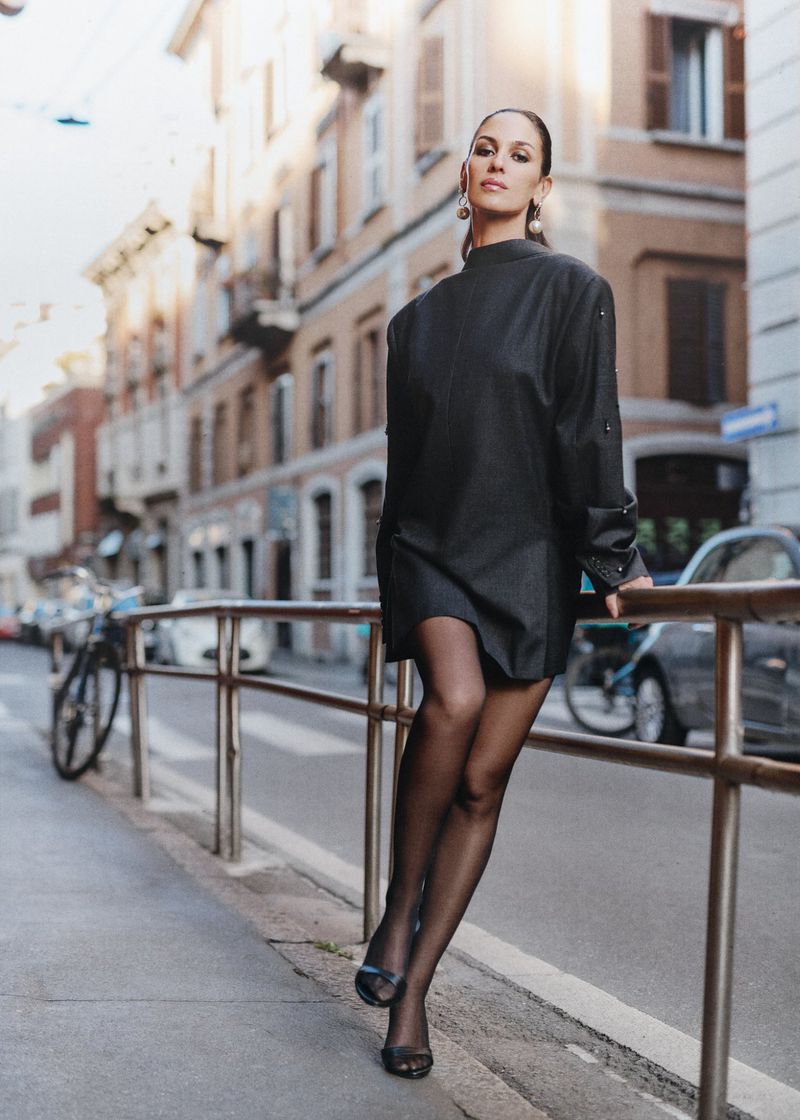 Woman in black dress and black tights posing on a city street near a metal railing, with buildings and parked vehicles in the background.