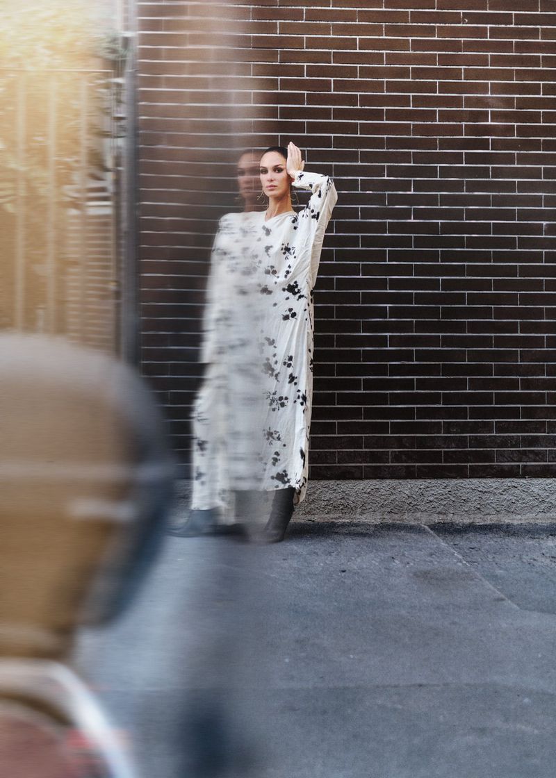 Woman in floral dress poses against a brick wall in an urban environment. She stands in blurred motion, with hand raised to her forehead.