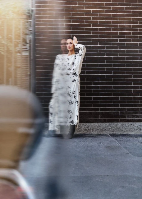 Woman in floral dress poses against a brick wall in an urban environment. She stands in blurred motion, with hand raised to her forehead.