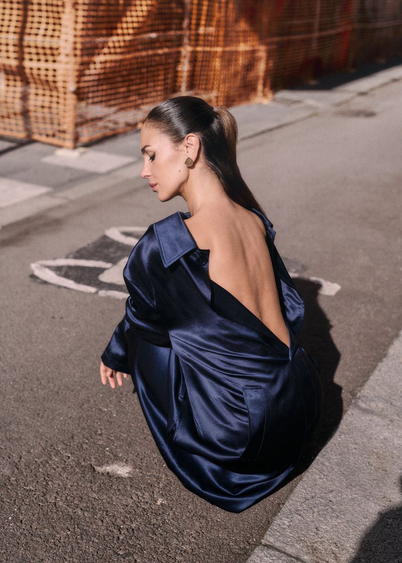 An adult female model with brown hair poses on a city street wearing a long-sleeve, backless, navy blue dress, with orange construction in the background.