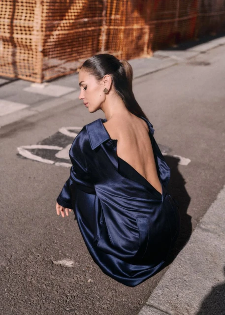 An adult female model with brown hair poses on a city street wearing a long-sleeve, backless, navy blue dress, with orange construction in the background.