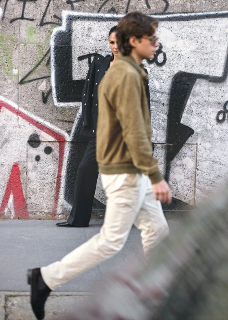 Two people walk past a graffiti wall. A woman in black stands with a man walking in front wearing a brown jacket and white pants.