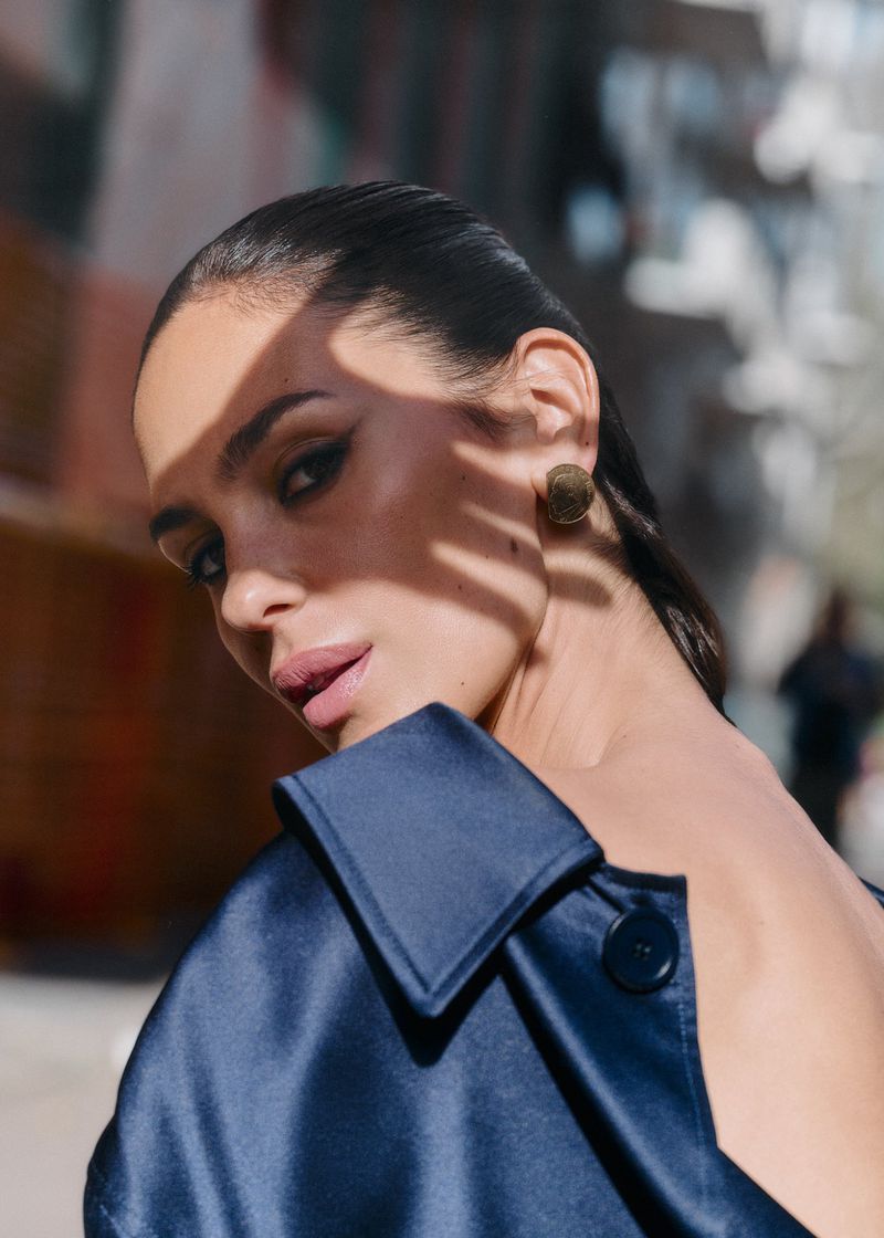 Close-up portrait of a woman with dark hair and satin coat, posing outdoors in natural light with building background and shadow patterns on face.