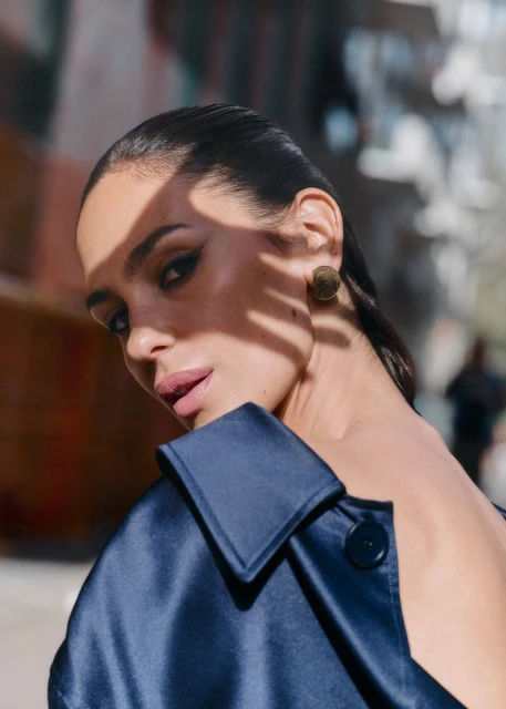 Close-up portrait of a woman with dark hair and satin coat, posing outdoors in natural light with building background and shadow patterns on face.
