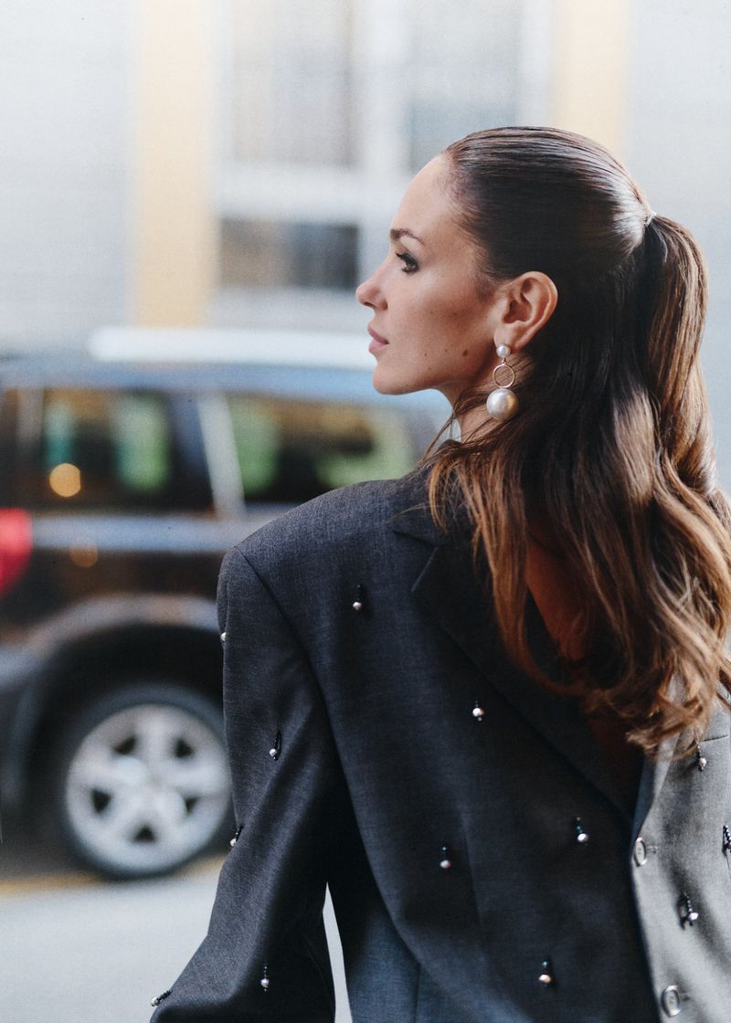 Adult woman in gray beaded blazer, side view. Ponytail hairstyle, statement earrings, and dark SUV in soft-focus background. 