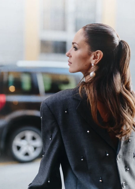 Adult woman in gray beaded blazer, side view. Ponytail hairstyle, statement earrings, and dark SUV in soft-focus background. 