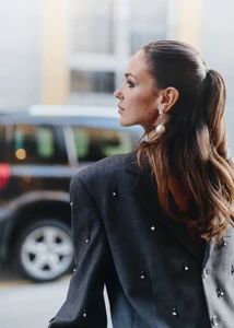 Woman in a pearl-embellished charcoal blazer and drop pearl earring facing in profile on a city street.