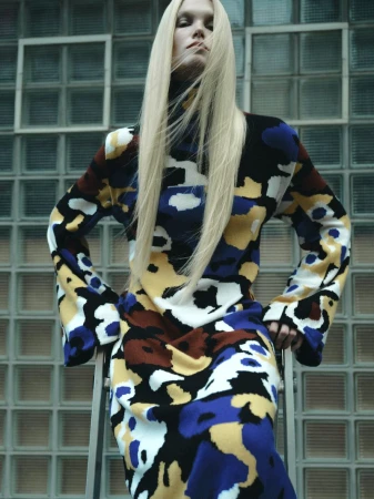 Model Sasha Dom with long straight blonde hair wearing a multi-colored abstract patterned knit dress in front of a glass block wall.