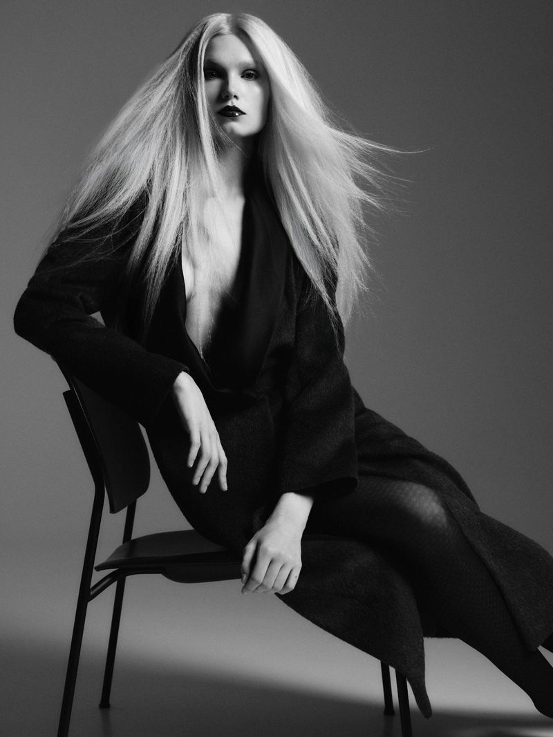 Model Sasha Dom seated on a dark chair with windblown long blonde hair wearing a black coat in a studio fashion portrait.