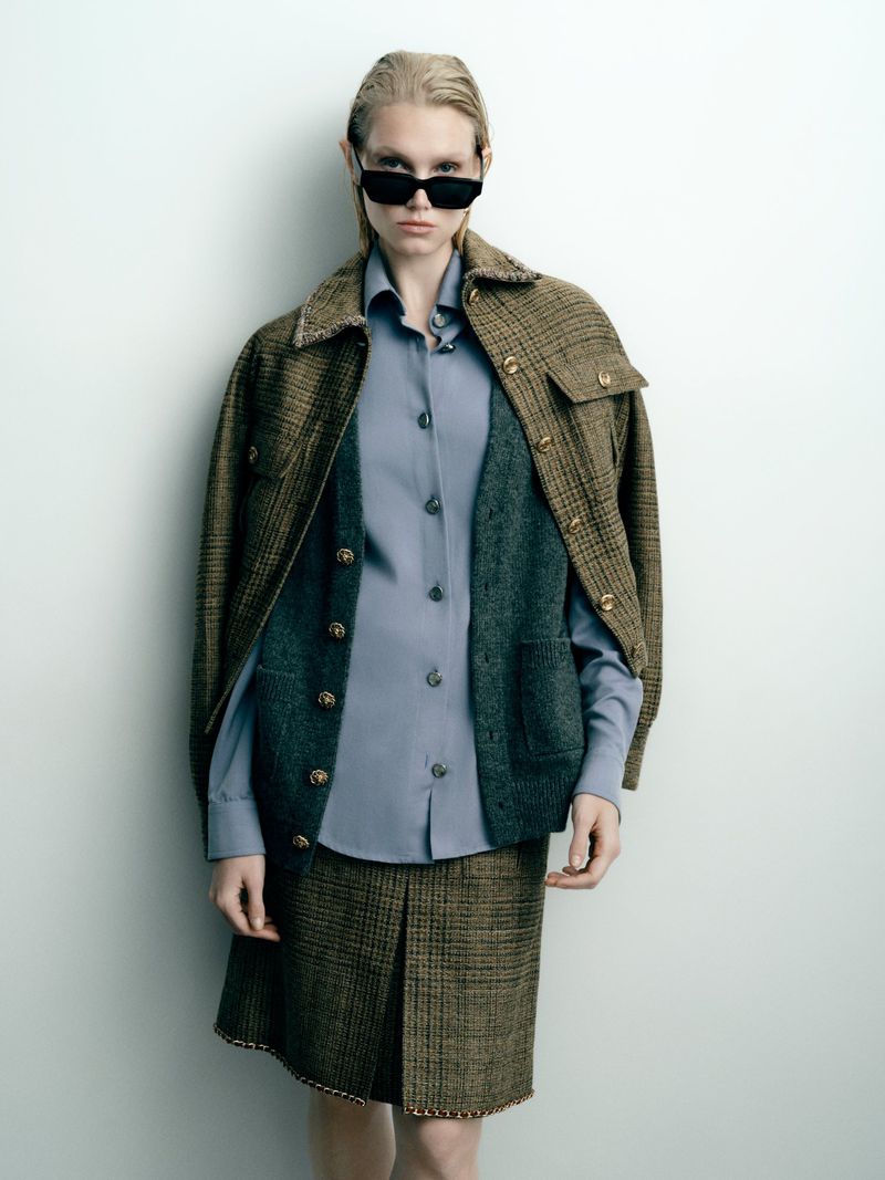 Sasha Dom poses in a layered tweed jacket, green knit cardigan, blue button-up shirt, and black sunglasses against a studio backdrop.