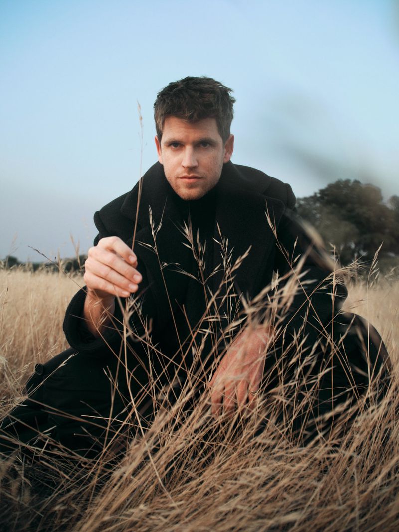 Miguel Bernardeau crouching in tall dry grass wearing a black coat and black turtleneck sweater.