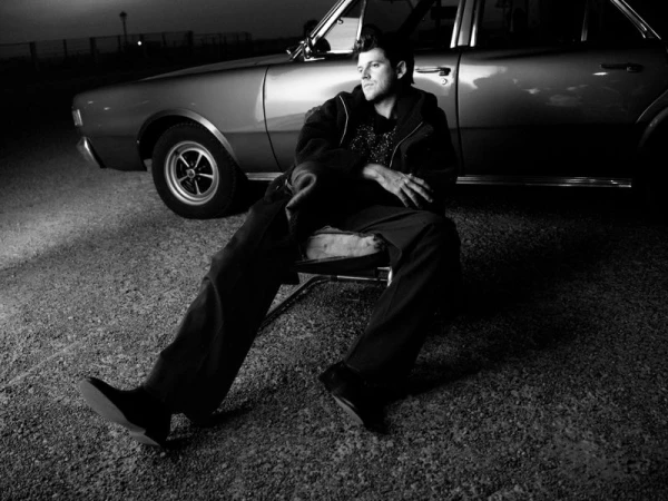 Monochrome editorial portrait of actor Miguel Bernardeau seated in a chair next to a vintage car on gravel at night.