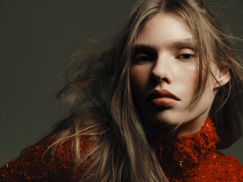 Close-up portrait of model Cara Schädel in a textured rust-orange high-neck sweater against a dark backdrop.