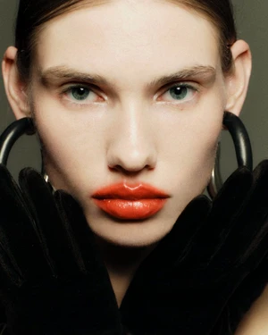 Close-up beauty portrait of a woman wearing glossy coral-red lipstick, oversized black hoop earrings, and black velvet gloves.