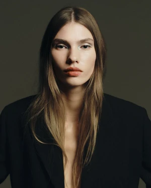 Model Cara Schädel looking directly into the camera wearing an open black blazer against a dark studio backdrop.