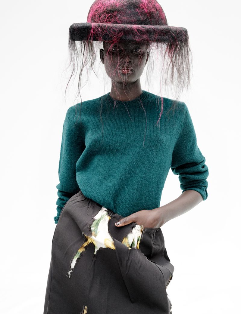 Model Aluel Keror wearing a green knit sweater and a fringed brimmed hat over her face against a bright white background.