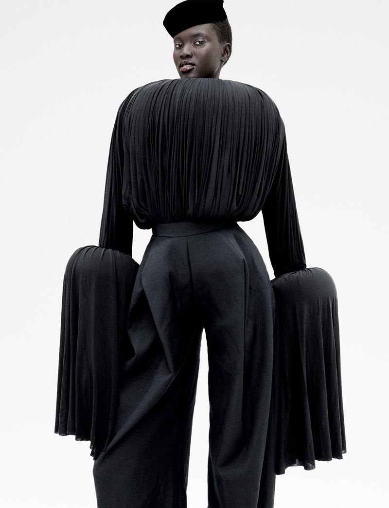 Model Aluel Keror wearing a sculptural black pleated top, wide-leg trousers, and a black hat against a white background.