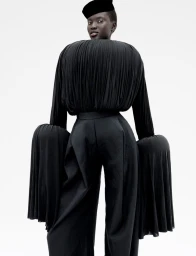 Model Aluel Keror wearing a sculptural black pleated top, wide-leg trousers, and a black hat against a white background.