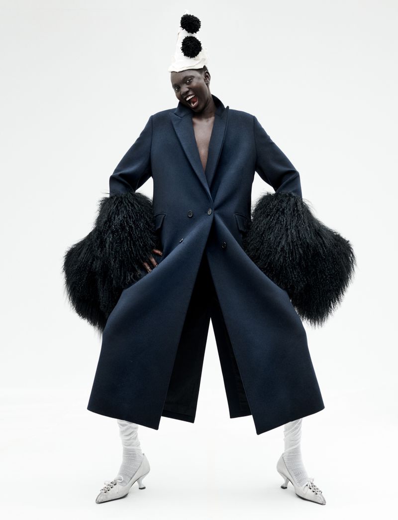 Model Aluel Keror poses in a dark navy coat with voluminous fur cuffs and a pompom party hat against a white seamless background.