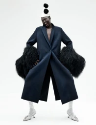 Model Aluel Keror poses in a dark navy coat with voluminous fur cuffs and a pompom party hat against a white seamless background.