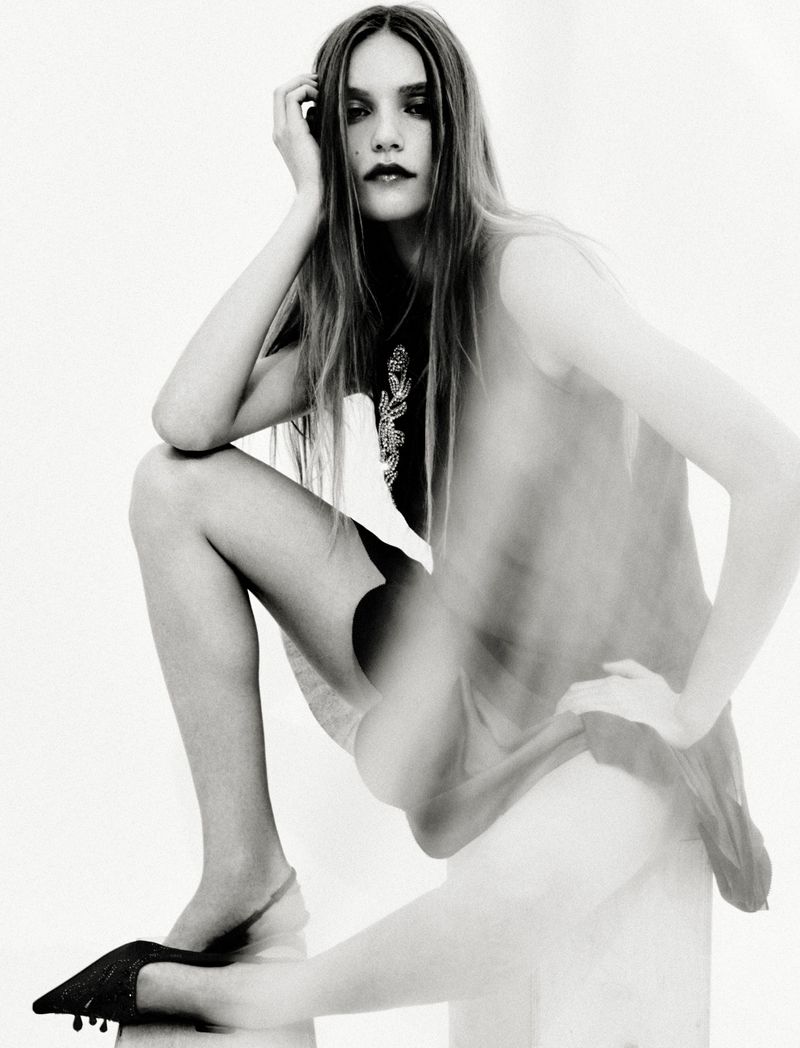Model Taylor D crouching in a sheer layered garment and black pointed heels against a stark white studio backdrop.