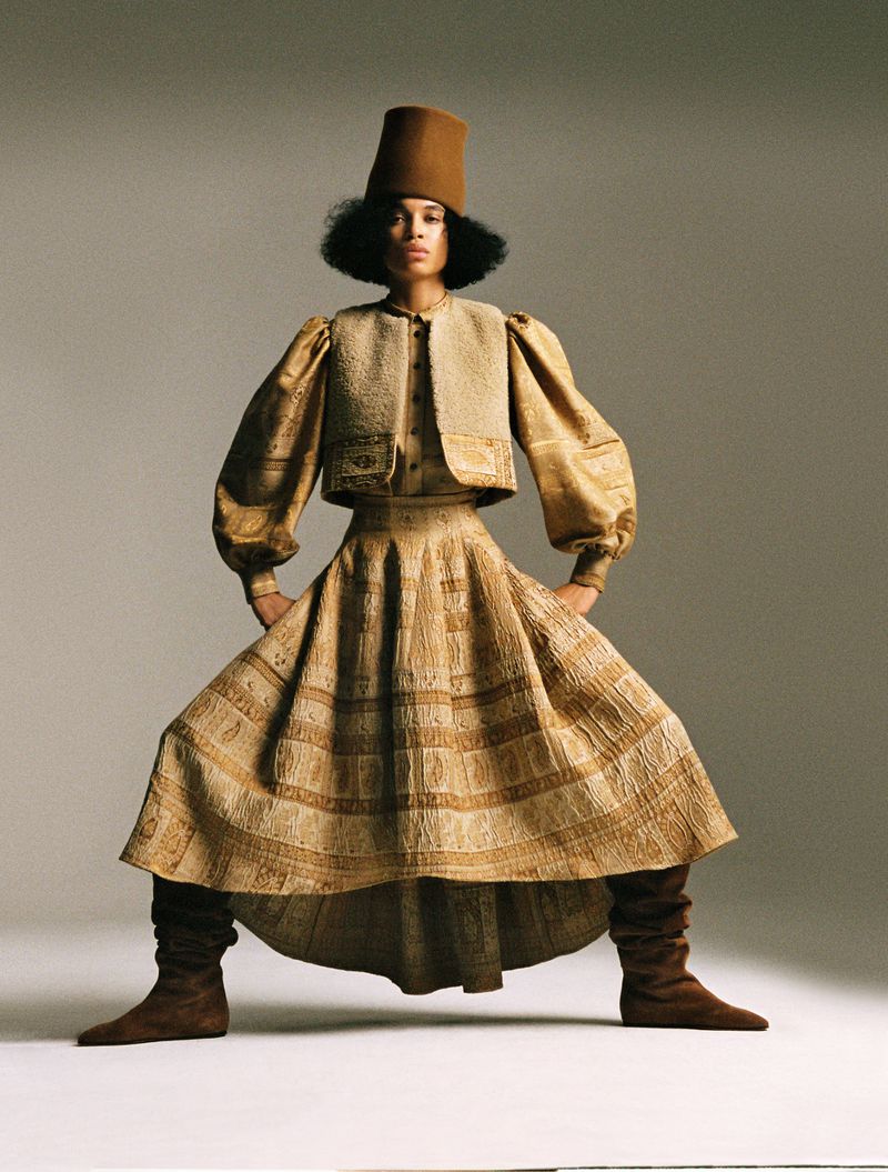 Model Lara Menezes in a wide stance wearing an embroidered tan skirt, cropped vest, tall brown hat, and slouch boots in a studio.