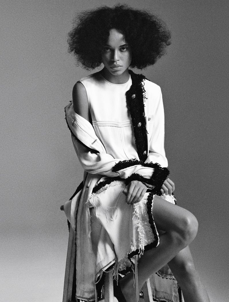 Black and white studio portrait of model Lara Menezes seated in a white frayed dress with dark trim against a seamless backdrop.