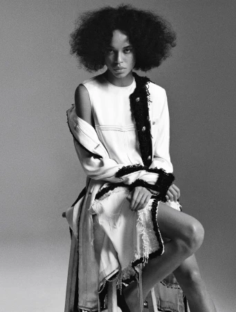 Black and white studio portrait of model Lara Menezes seated in a white frayed dress with dark trim against a seamless backdrop.