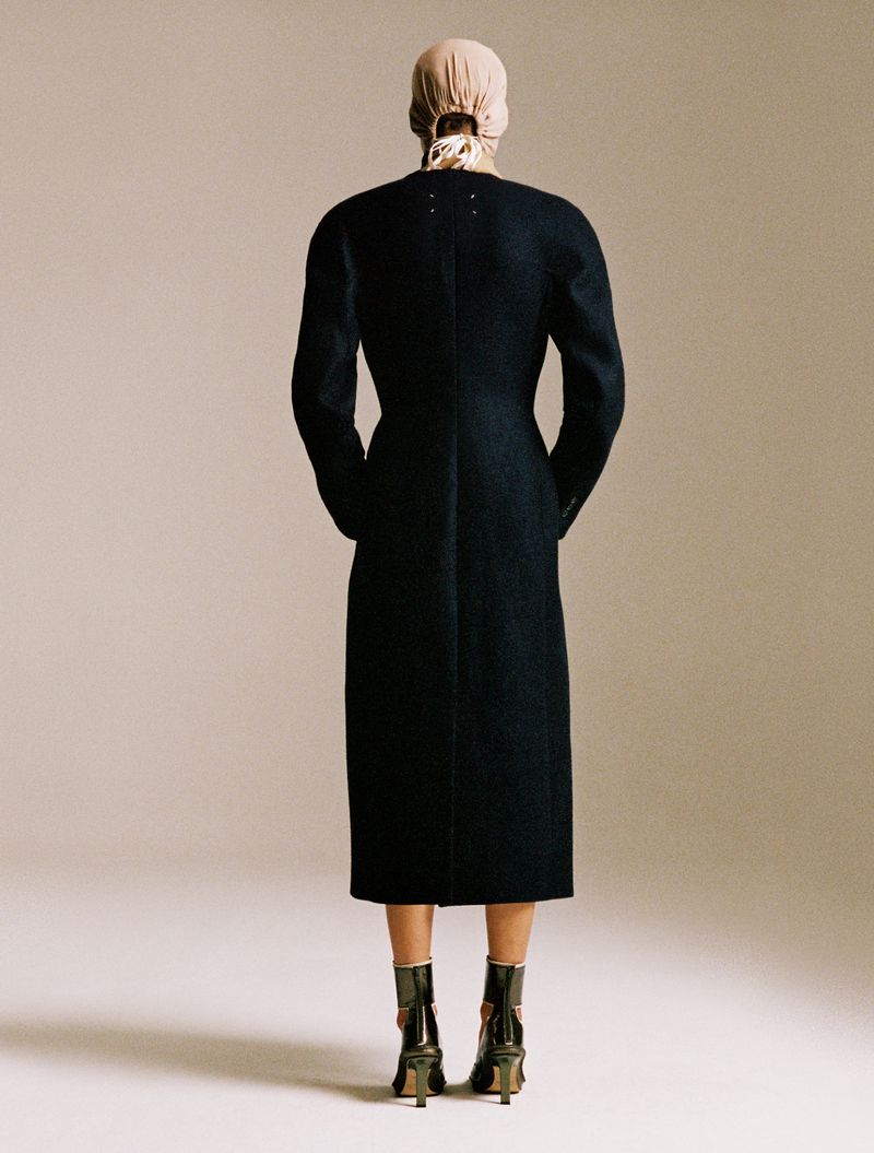 Model Lara Menezes viewed from behind wearing a tailored long black coat and head covering against a beige backdrop.