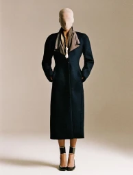Model standing in a structured dark long coat with a sheer mask over her head and face against a neutral studio backdrop.
