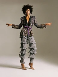 Model Lara Menezes poses in a metallic embroidered jacket, purple sash, and tiered pleated grey trousers against a studio backdrop.