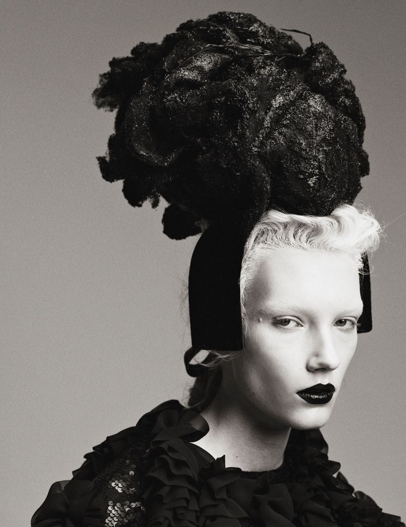 Model Ivy Stewart wearing an avant-garde black sculpted headpiece, dark lipstick, and ruffled collar against a grey studio backdrop.