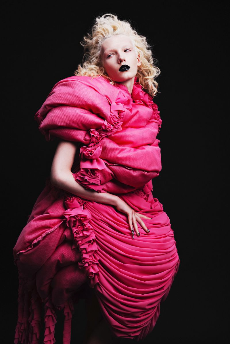 Model Ivy Stewart posing in a voluminous sculptural hot pink ruffled dress against a black background.
