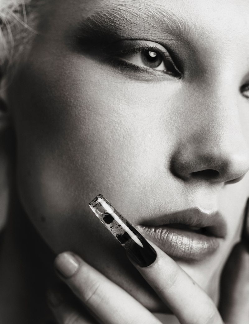 Monochrome close-up portrait of model Ivy Stewart with dark eye makeup touching her face with a sculptural black-tipped nail.