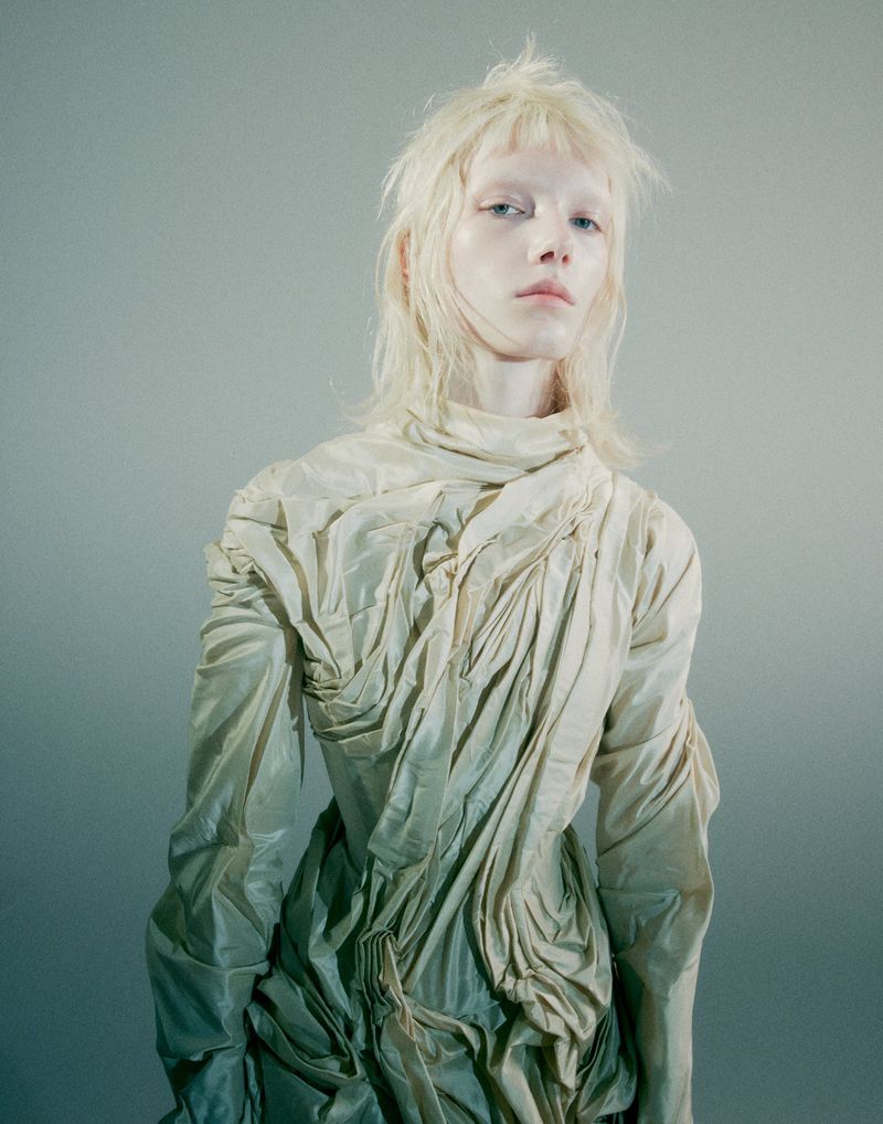 Model Ivy Stewart posing in a high-neck ruched cream garment with shaggy blonde hair against a soft grey backdrop.