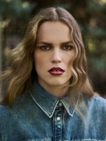 Portrait of model Mathilde Lakjaer wearing a washed denim button-up shirt with wavy hair and bold dark berry lipstick.