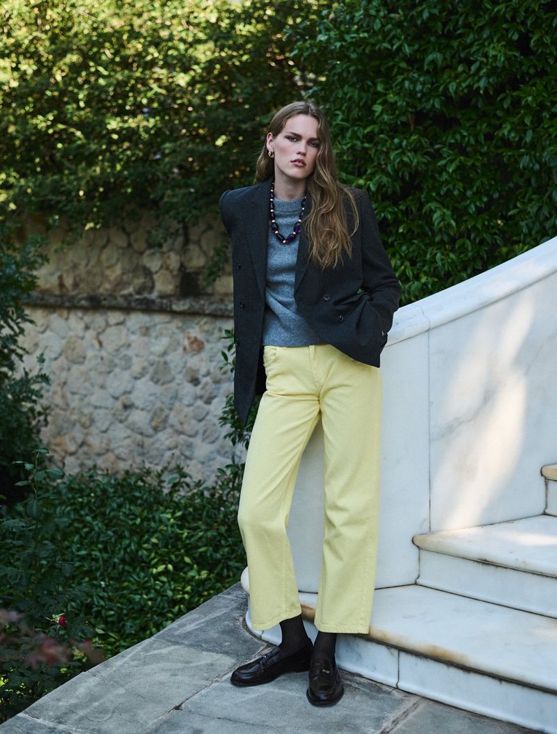 Model Mathilde Lakjaer in a dark charcoal blazer and pastel yellow trousers standing on outdoor stone steps.