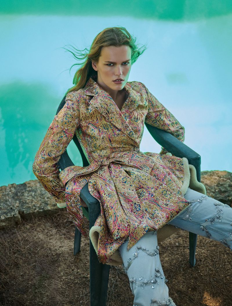 Mathilde Lakjaer seated in a green plastic chair by turquoise water wearing a brocade coat and embellished light blue jeans.