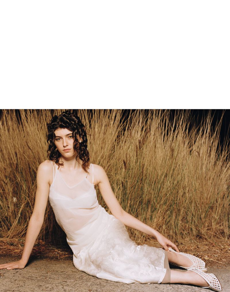 Young woman in a white dress sits on the ground in front of tall grass, looking at the camera with a neutral facial expression.