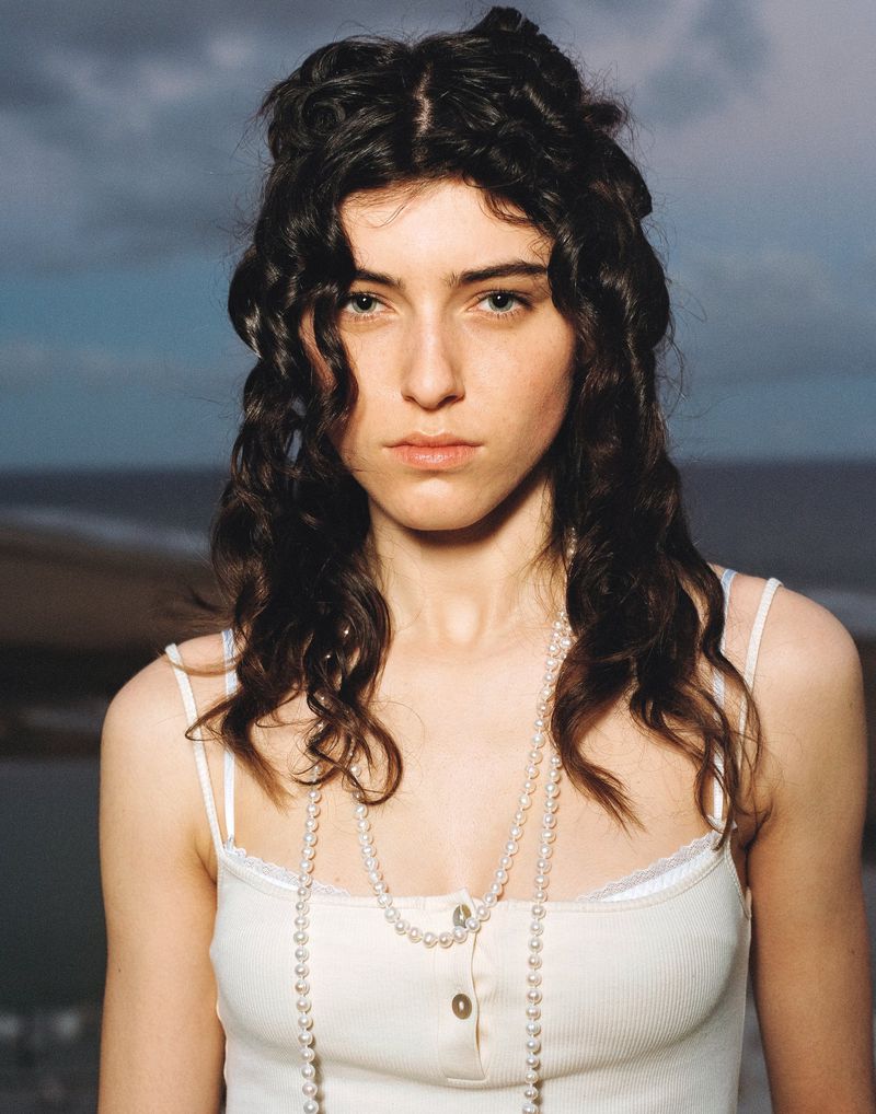 Portrait of a young adult female with long dark curly hair wearing a white top and pearls against an ocean and cloudy sky.