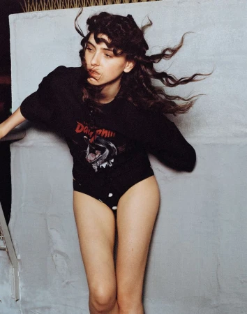 Brunette woman with flowing hair and a printed black shirt poses against a gray wall, capturing a casual yet stylish moment.