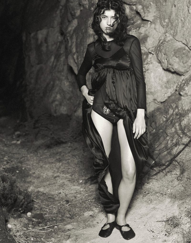 Monochrome portrait of a brunette woman in a black dress standing near a rock wall, looking at the camera in a fashion editorial style.