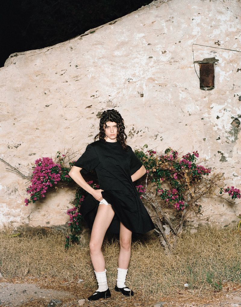 A young woman poses in a black dress outside, standing in a grassy field with a weathered building and pink flowering bush behind her.