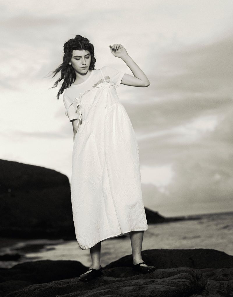 Young woman in a white dress posing on rocks near the sea with a cloudy sky, creating a timeless, monochrome aesthetic.