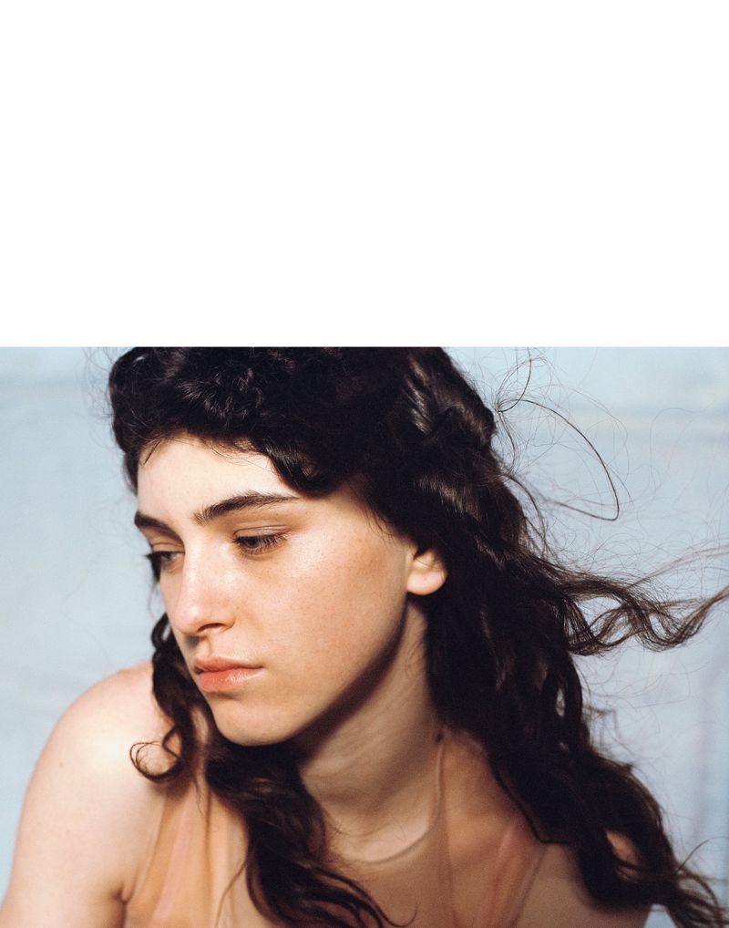 Portrait of a young woman with dark wavy hair, looking down in soft lighting. The image captures a serene and intimate moment.