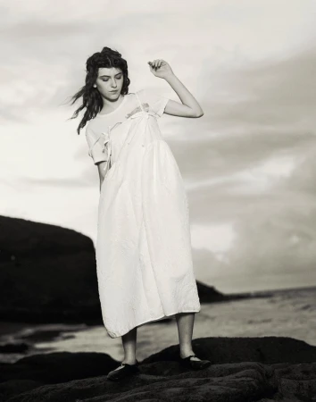 Model Margaux Martin wearing a white layered dress while standing on dark coastal rocks overlooking the ocean under an overcast sky.