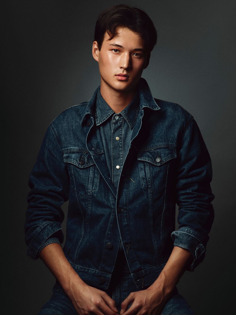 A young man in a dark blue denim jacket and matching shirt looking forward in a studio portrait.