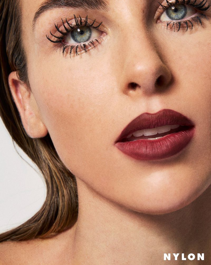 Close-up beauty portrait of a woman with bold matte burgundy lipstick and dramatic spidery eyelashes against a light background.