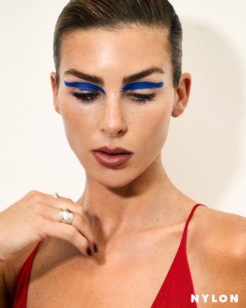 Portrait of a woman with bold graphic royal blue eyeshadow wearing a red spaghetti strap top against an off-white background.