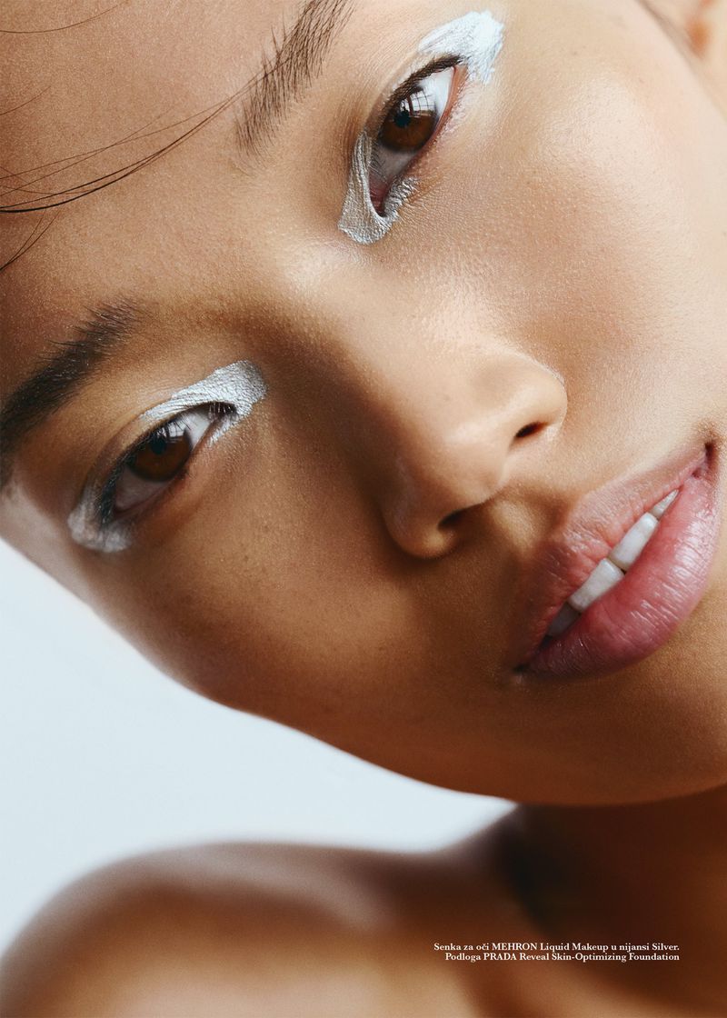Close-up portrait of an Asian woman with graphic metallic silver eye makeup and glowing skin, tilting her head against a white background.